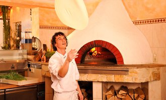 Ein Pizzabäcker wirft einen Pizzateig in die Luft. Im Hintergrund ist ein traditioneller Holzofen zu sehen. | © Ristorante Trentino | Michael Monschau