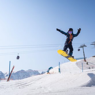 A snowboarder jumps over a snow hill in front of a clear mountain landscape. In the background, ski lifts and other skiers can be seen. | © OBERSTDORF · KLEINWALSERTAL BERGBAHNEN