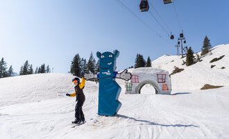 Ein Skifahrer steht auf schneeweißer Piste neben einem blauen Bären und einem architektonischen Element. Im Hintergrund sind Lifte und verschneite Bäume zu sehen. | © OBERSTDORF · KLEINWALSERTAL BERGBAHNEN