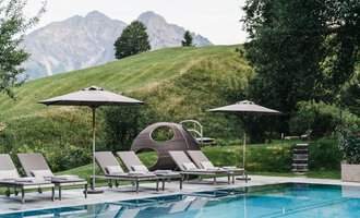 A pool area with loungers and sun umbrellas. In the background, gentle hills and mountains can be seen.
