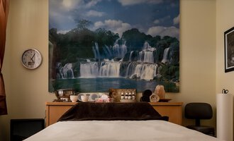 Ein gemütlicher Raum mit einem Bett und einer großen Wandmalerei von Wasserfällen. Auf dem Tisch sind verschiedene Dekorationsgegenstände und eine Uhr zu sehen. | © IFA Hotel Alpenhof Wildental | Andreas Froschauer