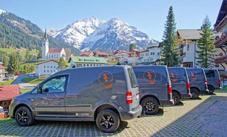 Four delivery vans are parked on a paved area with a picturesque mountain backdrop. In the background, green meadows, houses, and a church are visible. | © Der Elektriker Markus Leitgeb GmbH