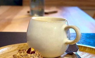 A cup of coffee on a wooden board next to a cookie. In the background, there is a vase with colorful flowers visible. | © Einkehr Guntschau