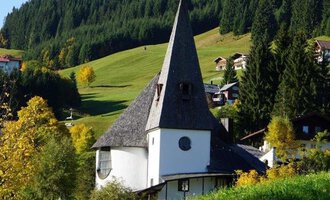 Eine malerische Kirche mit einem spitzen Dach in einer hügeligen Landschaft. Im Hintergrund sind grüne Wälder und einige kleine Gebäude zu sehen. | © Ev. Kirche Kleinwalsertal