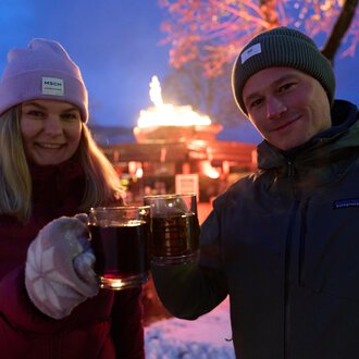 Two people are clinking glasses and smiling. In the background, a festive atmosphere with a fire pit and seasonal lights can be seen.
