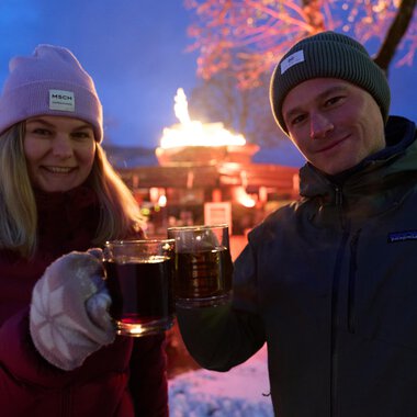 Two people are clinking glasses and smiling. In the background, a festive atmosphere with a fire pit and seasonal lights can be seen.