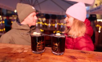 Two cups of hot drinks are on a wooden table. In the background, a man and a woman are talking, surrounded by festive lighting.