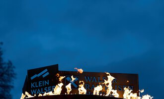 Ein Schild mit der Aufschrift "Klein Walsertal" steht in Flammen vor einem dunklen Himmel. Die Feuer leuchten hell und schaffen eine dramatische Stimmung.
