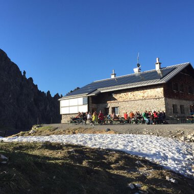 A mountain hut in the Alps surrounded by snow-covered ground. Many people are sitting outside enjoying the sun under a blue sky. | © Kleinwalsertal Tourismus