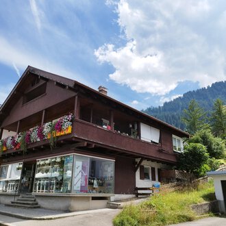 Ein charmantes Haus mit einem blühenden Balkon in den Bergen. Die Umgebung ist grün und die Wolkenhimmel sorgt für eine schöne Atmosphäre. | © Kleinwalsertal Tourismus | N. Lughammer