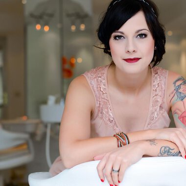A woman with tattoos is sitting smiling in a modern room. She is wearing an elegant pink dress and has a chic look. | © Fräulein Sugar Coiffeur | Sandra Bromm