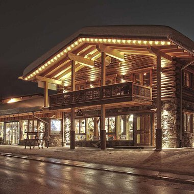 A cozy wooden building with warm light at night. The snow-covered ground and stylish design create an inviting atmosphere. | © Fun-Alp
