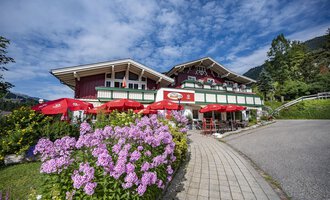 A picturesque alpine house with red sunshades and colorful flowers. The sky is blue with some clouds. | © Gasthof Alpenblick | Theodor Pinn