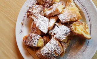 Ein Teller mit gebackenem Kuchen, bestäubt mit Puderzucker. Die Stücke sind goldbraun und appetitlich angerichtet. | © Gasthof Café Alpenwald | Thorsten Alpenwald