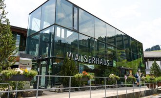 Ein modernes Gebäude mit Glasfassade steht im Vordergrund. Umgeben von Bäumen und Blumen, vermittelt es einen einladenden Eindruck. | © Kleinwalsertal Tourismus | Oliver Farys