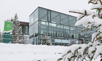 Ein modernes Gebäude mit großen Glasfenstern im Schnee. Umgeben von verschneiten Bäumen zeigt es eine winterliche Atmosphäre. | © Kleinwalsertal Tourismus | Oliver Farys