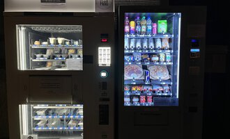Zwei Verkaufsautomaten stehen nebeneinander. Der linke Automat bietet gefrorene Lebensmittel, während der rechte verschiedene Getränke und Snacks enthält.
