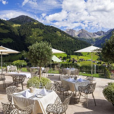 A stylish outdoor restaurant with a view of the mountains. There are elegant tables with umbrellas and green plants. | © Hallers Geniesserhotel | Dietmar Walser