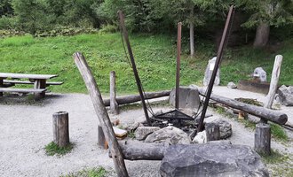 Ein gemütlicher Grillplatz im Freien mit einem großen Feuerkreis aus Steinen und Holz. Im Hintergrund steht eine Picknickbank auf einer grünen Wiese. | © Kleinwalsertal Tourismus | Veronika Senn