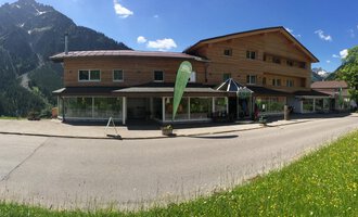 Ein modernes Gebäude in den Bergen, umgeben von grüner Wiese und blauen Himmel. Im Vordergrund ist eine gepflasterte Straße sichtbar. | © Sport Hilbrand GmbH