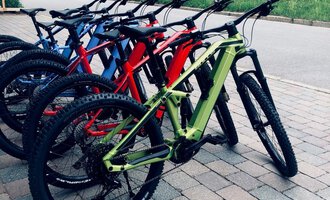 A row of colorful mountain bikes stands on a paved path. In the background, a wooden house and a green meadow can be seen. | © Sport Hilbrand GmbH