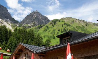 A cozy restaurant in the mountains with terraces and red umbrellas. Majestic mountains and a blue sky can be seen in the background. | © Hintere Gemstelhütte | Frank Drechsel
