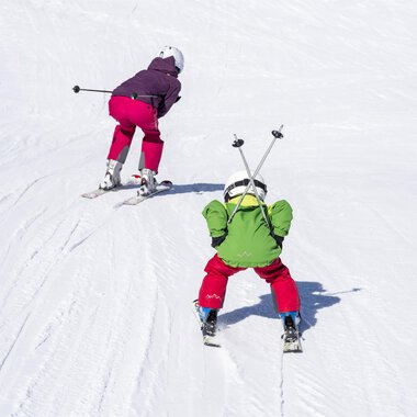 Two children are skiing on a snowy slope. One child is wearing a green suit and the other a purple suit. | © Kleinwalsertal Tourismus | Dominik Berchtold
