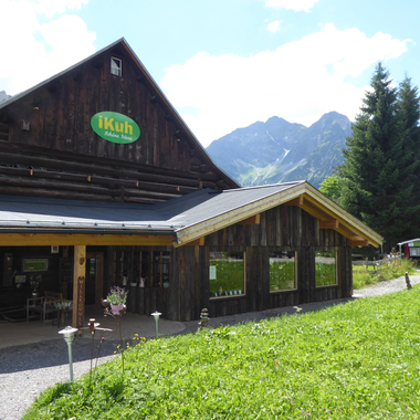 A rustic wooden building with a green sign. In the background, there are tall mountains and a blue sky. | © iKuh Schöne Ideen | Daniela Neumann