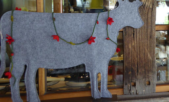 A gray fabric cow, decorated with red flowers. It stands in front of a wooden shelf in a rustic setting. | © iKuh Schöne Ideen | Daniela Neumann