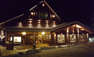 Der Laden iKuh Schöne Ideen in einem gemütlichen Holzhaus bei Nacht, beleuchtet von warmem Licht. Es ist von Schnee umgeben und strahlt eine gemütliche Atmosphäre aus.