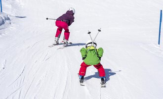 Two children are skiing on a snowy slope. One child is wearing a green jacket and the other a purple jacket. | © Kleinwalsertal Tourismus | Dominik Berchtold