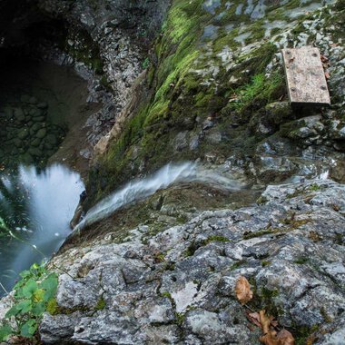 Eine felsige Klippe mit fließendem Wasser, das in einen tiefen, dunklen Pool mündet. Grüne Moose und Pflanzen wachsen zwischen den Steinen. | © Kleinwalsertal Tourismus | Andre Tappe