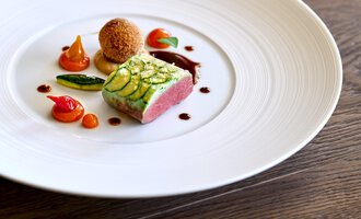 A stylish dish on a white plate, consisting of meat with zucchini, surrounded by colorful vegetables and a sauce. The presentation is elegant and appealing.