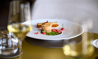 An artfully arranged plate with a fine dish, surrounded by decorative berries and a green sauce. In the background, wine glasses can be seen.