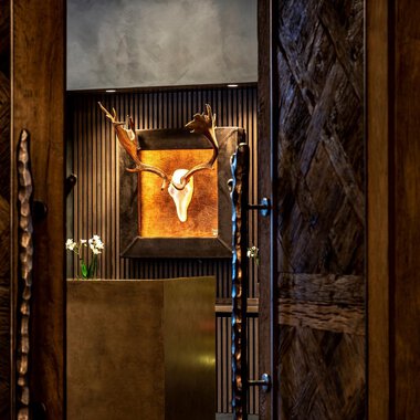 A stylish entrance area with wooden walls and a decorative antler. In the background, a piece of artwork featuring an animal head can be seen. | © Travel Charme Ifen Hotel