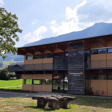 A modern building with large windows and wooden cladding. In the foreground, there is a stone table on a green meadow with mountains in the background. | © Kleinwalsertal Tourismus | Veronika Senn