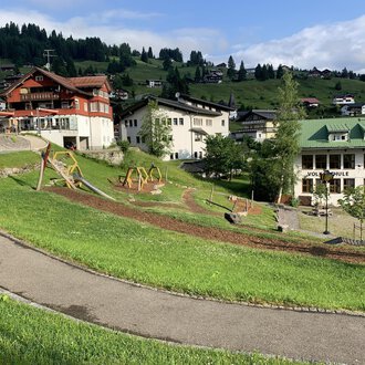 A picturesque landscape with green meadows and playgrounds. In the background, some chalet-like buildings and mountains can be seen. | © Kleinwalsertal Tourismus