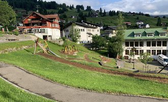 A picturesque landscape with green meadows and playgrounds. In the background, some chalet-like buildings and mountains can be seen. | © Kleinwalsertal Tourismus