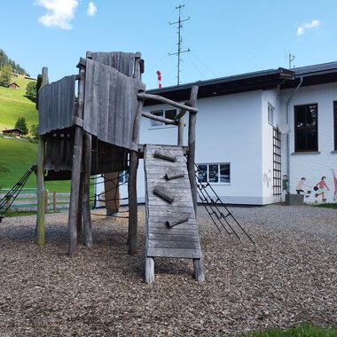 A playground with a wooden climbing structure and a slide. In the background, you can see a house and green hills. | © Kleinwalsertal Tourismus | N. Lughammer