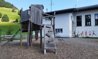 A playground with a wooden climbing structure and a slide. In the background, you can see a house and green hills. | © Kleinwalsertal Tourismus | N. Lughammer