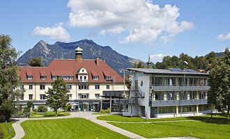 A charming hotel building with a red roof and modern architecture. In the background, mountains and a blue sky can be seen. | © Klinikverbund Allgäu