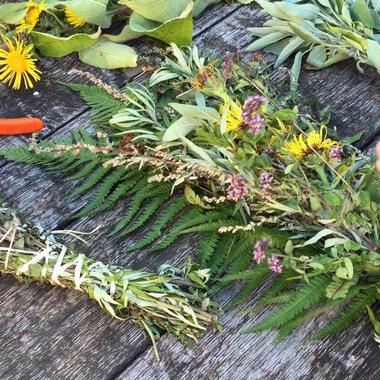 Ein Tisch mit frischen Kräutern und bunten Blumen. Jemand bindet einen Kräuterkranz mit einer Schere daneben. | © Kleinwalsertal Tourismus | Lydia Fritz-Ilg