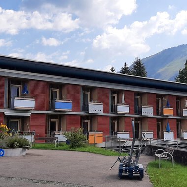 Ein modernes Gebäude mit mehreren Etagen und Balkonen. Im Hintergrund sind grüne Hügel und ein blauer Himmel zu sehen. | © Kleinwalsertal Tourismus | Veronika Senn