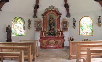 A small chapel with wooden bench rows and colorful windows. In the foreground stands an ornate altar with a statue. | © Stefan Heim