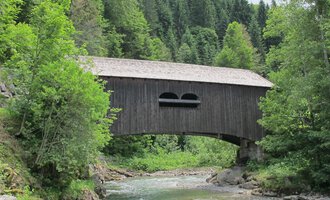 Eine alte Holzbrücke über einen klaren Fluss, umgeben von grünen Bäumen. Im Hintergrund erheben sich bewaldete Hügel. | © Stefan Heim