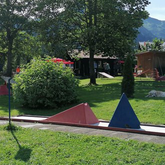 A mini golf course with colorful obstacles in a green environment. In the background, trees and part of a building can be seen.