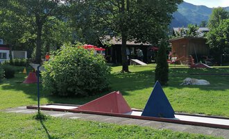 A mini golf course with colorful obstacles in a green environment. In the background, trees and part of a building can be seen.