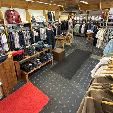 A fashionable clothing store with a variety of shirts and sweaters. The shelves are neatly organized, and the floor is covered with a red carpet as well as a patterned rug. | © Moosbrugger Mode e.U.
