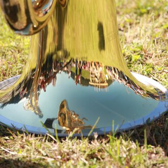 A shiny trumpet standing on the grass. Trees and people can be seen in the reflecting metal. | © Kleinwalsertal Tourismus | Frank Drechsel