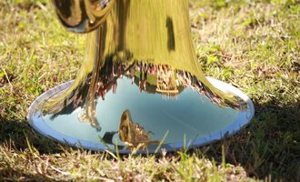 A shiny trumpet standing on the grass. Trees and people can be seen in the reflecting metal. | © Kleinwalsertal Tourismus | Frank Drechsel